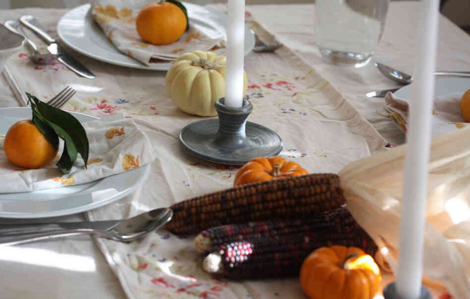 A dining table set for the holidays with linens, candlesticks, clementines and gourds.