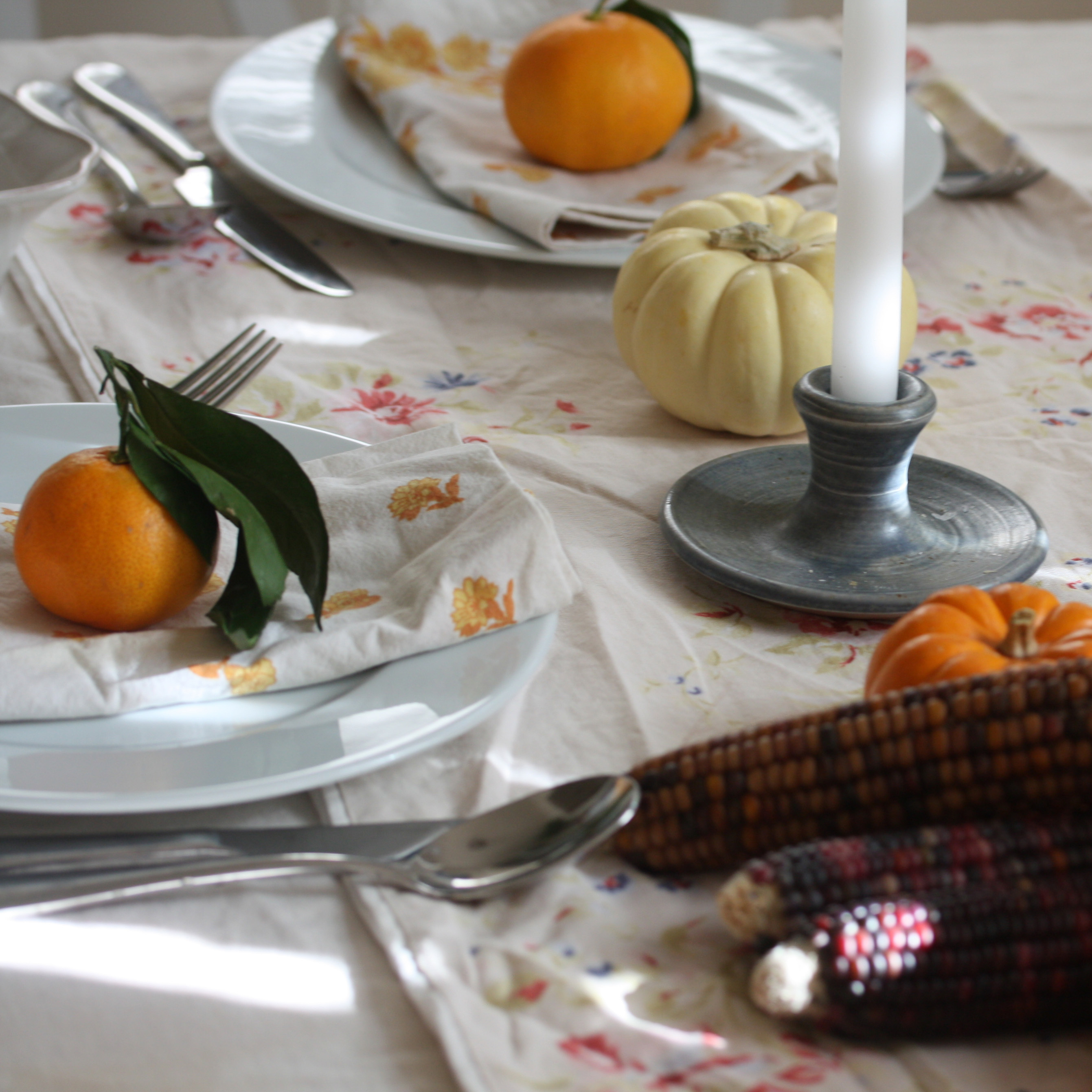 A dining table set for the holidays with linens, candlesticks, clementines and gourds.