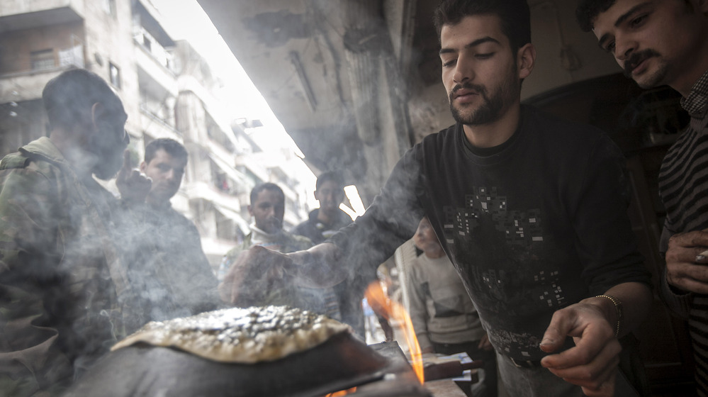 A man makes bread as residents, background, stand in line in front of a bakery during heavy fighting between Free Syrian Army fighters and government forces in Aleppo, Syria, on Dec. 4, 2012.