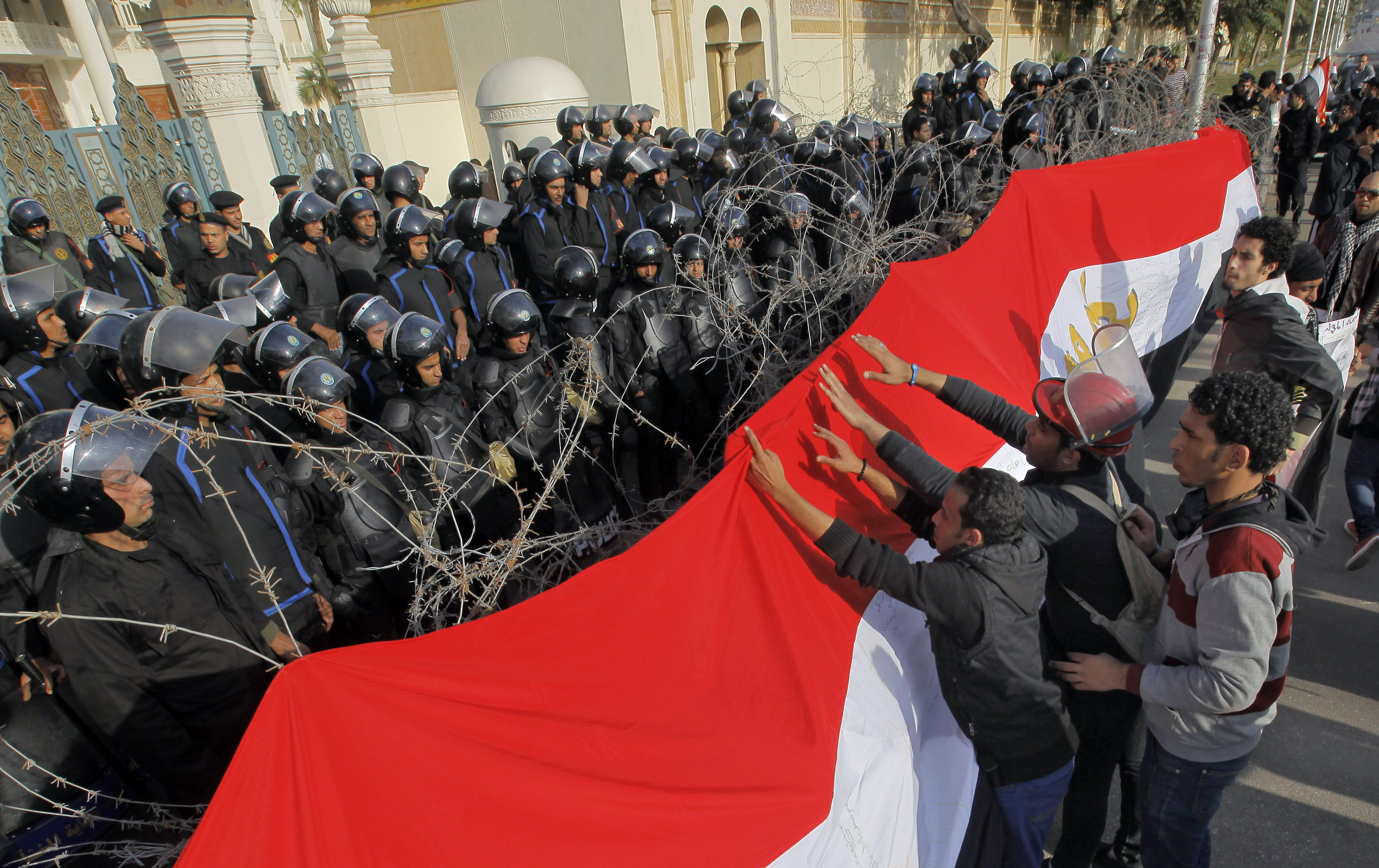Egyptian protesters hang a giant banner in the colors of Egypt's national flag on barbed wires in front of anti-riot soldiers at the entrance to the presidential palace in Cairo, Egypt, Friday, Jan. 25, 2013. 