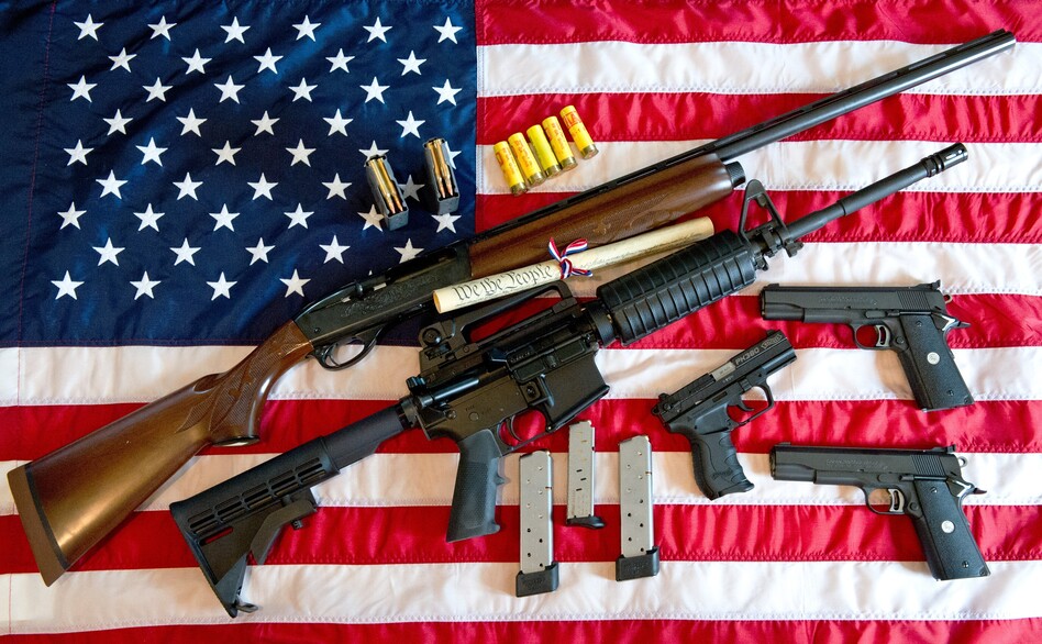 A photo illustration showing a Remington 20-gauge semi-automatic shotgun, a Colt AR-15 semi-automatic rifle, a Colt .45 semi-auto handgun, a Walther PK380 semi-auto handgun and various ammunition clips with a copy of the US Constitution on top of the American flag.