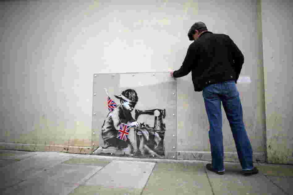 A man inspects a plastic cover placed over an artwork attributed to Banksy in London, England. The stencilled image depicts a poor child making Union Jack flags on a sewing machine and is located on the wall of a Poundland discount shop in the Wood Green area of north London.
