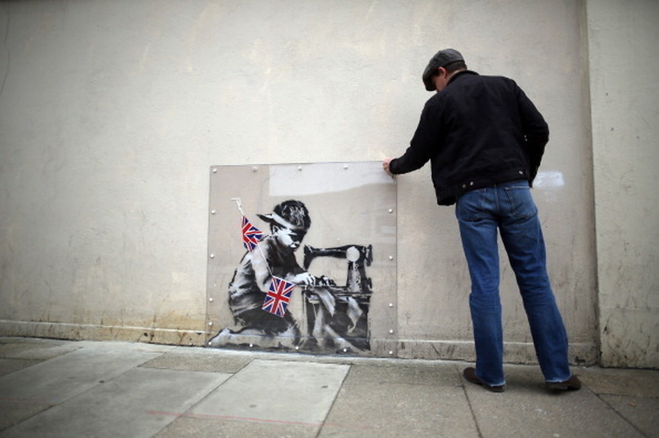 A man inspects a plastic cover placed over an artwork attributed to Banksy in London. The stencilled image depicts a poor child making Union Jack flags on a sewing machine and is located on the wall of a Poundland discount shop in the Wood Green area of north London.