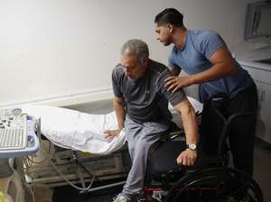 Joel Ramirez is helped onto a bed to receive an ultrasound with the aid of Francisco Guardado, his home health care giver, during a visit to the hospital in Rialto, Calif.