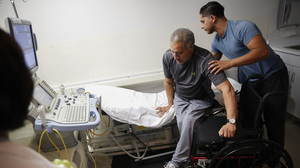 Joel Ramirez is helped onto a bed to receive an ultrasound with the aid of Francisco Guardado, his home health care giver, during a visit to the hospital in Rialto, Calif.