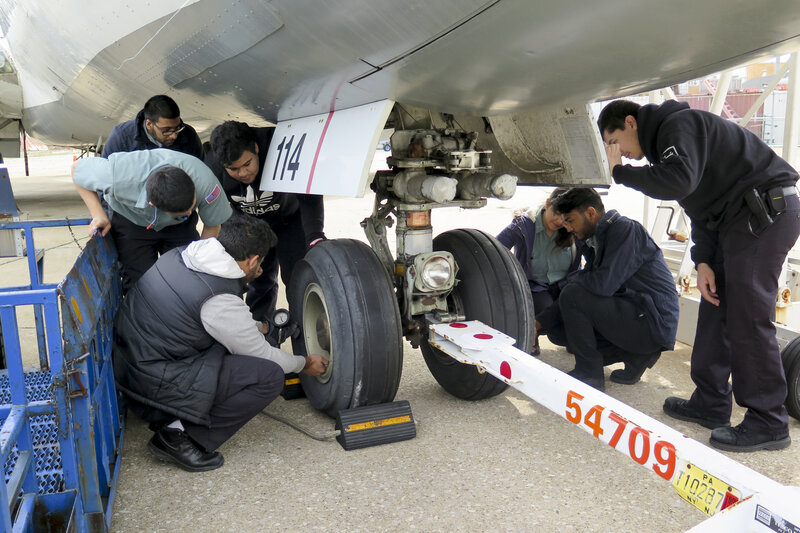 Students gather under the wheel carriage to practice checking the tire pressure. "It's scary business," warns their teacher, Mike Fisher. "If somebody tried to put the pressure from the bottle straight into the tire, they'd blow the tire up." Students gather under the wheel carriage to practice checking the tire pressure. "It's scary business," warns their teacher, Mike Fisher. "If somebody tried to put the pressure from the bottle straight into the tire, they'd blow the tire up."