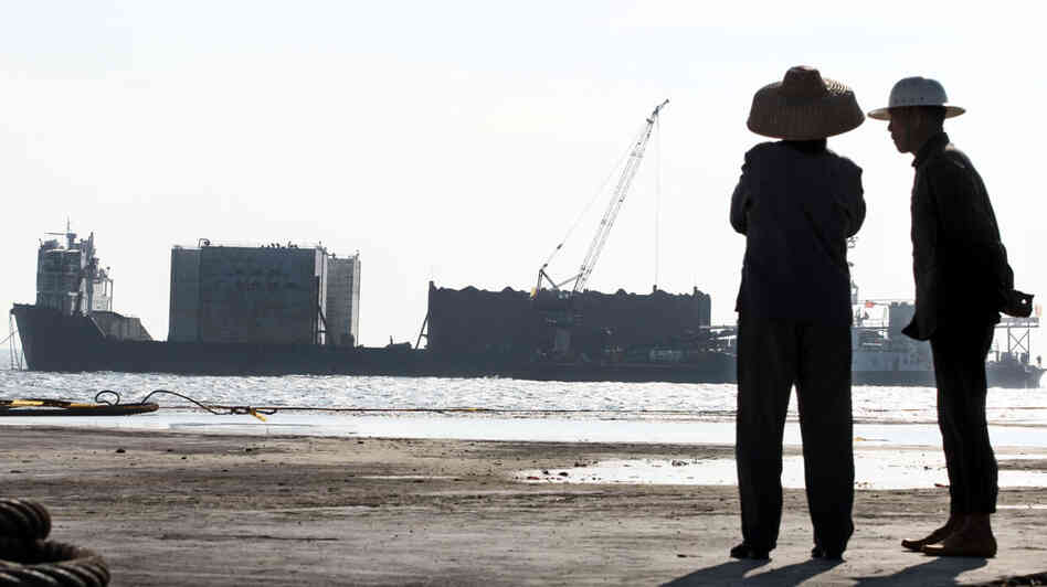 Fishermen watch as a barge carries the shipwreck