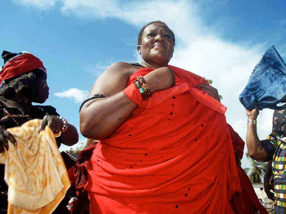 King Peggy, who is wearing traditional red funeral fabric, is being fanned by attendants. 