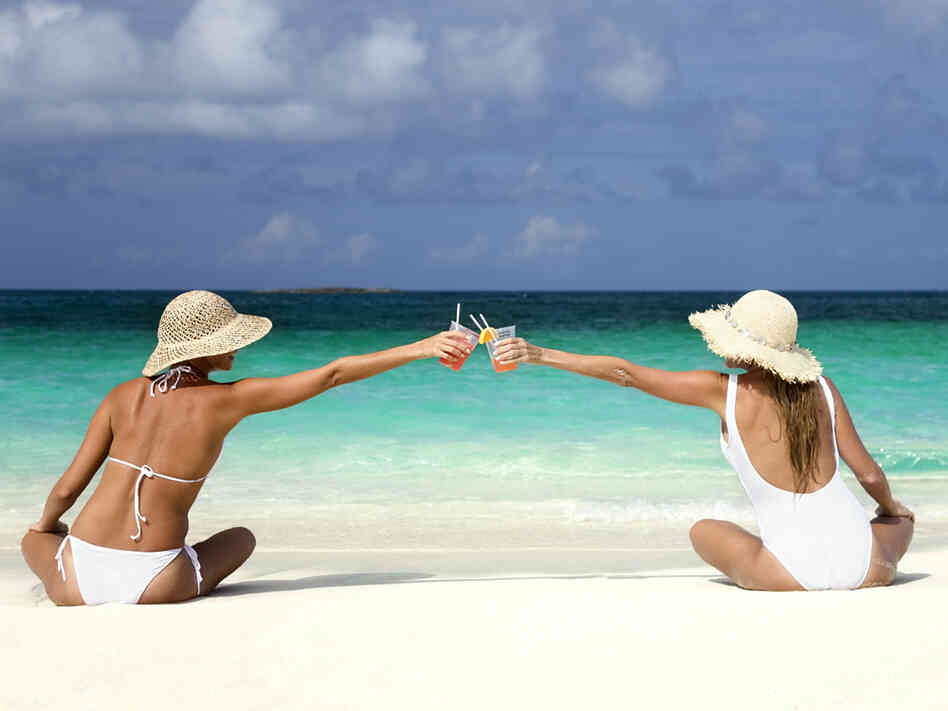 Two women share cocktails on a beach.