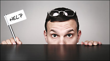 a man peeks over a desk, holding up a sign that says 'help'