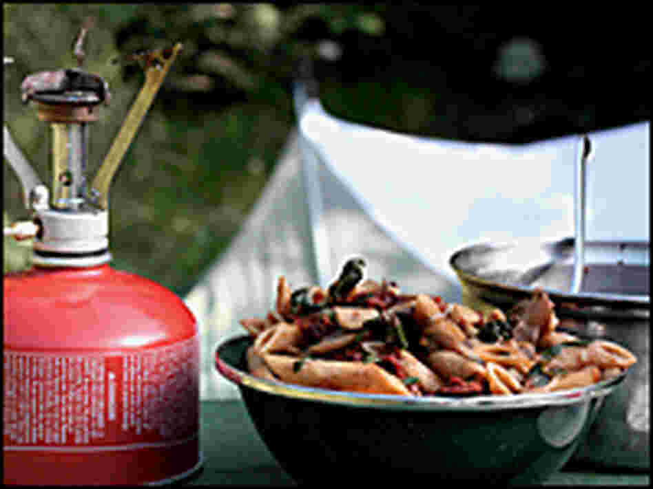 A bowl of penne pasta with vegetables sits on a picnic table in front of a tent
