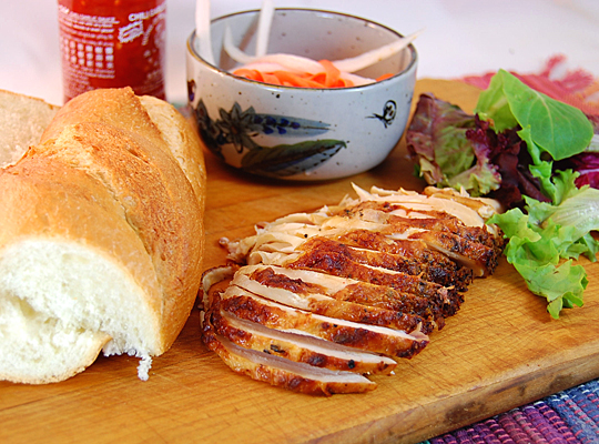 Fixings for Banh Mi -- crusty bread, sliced meat, pickled radish, chili sauce -- on a cutting board
