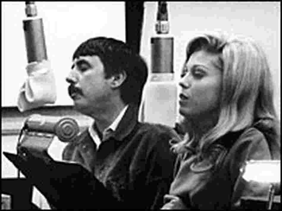 Lee Hazlewood and Nancy Sinatra, seated at music stands and microphones, sing in a recording studio