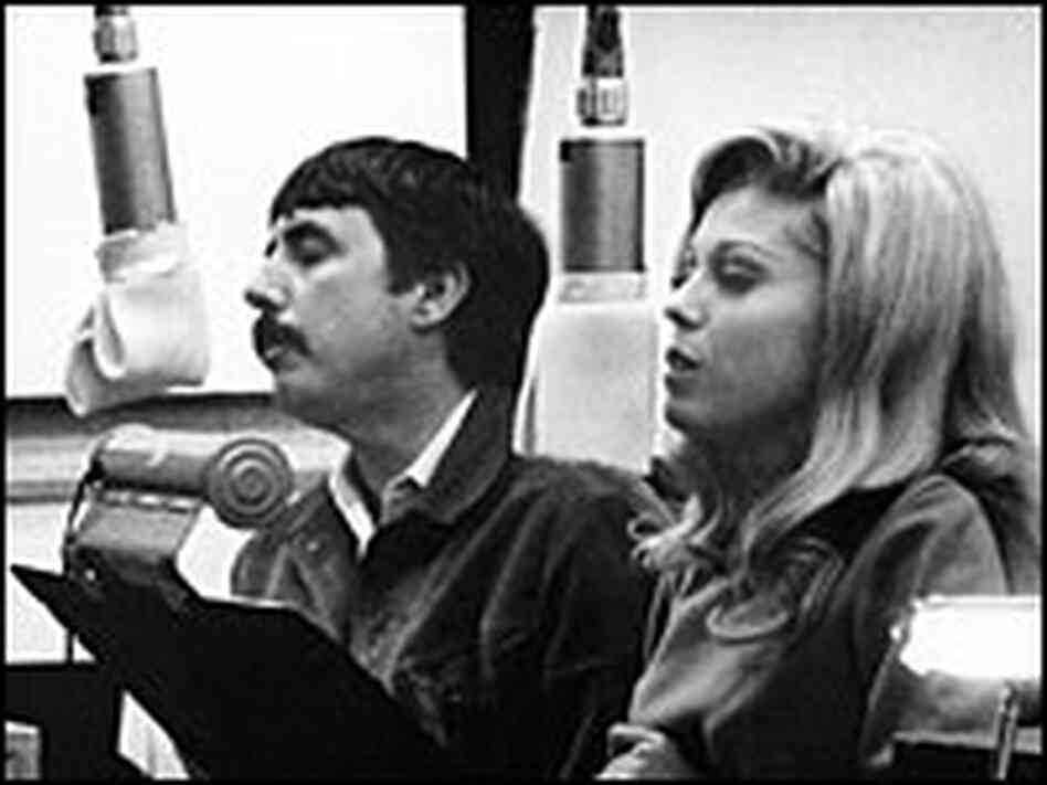 Lee Hazlewood and Nancy Sinatra, seated at music stands and microphones, sing in a recording studio