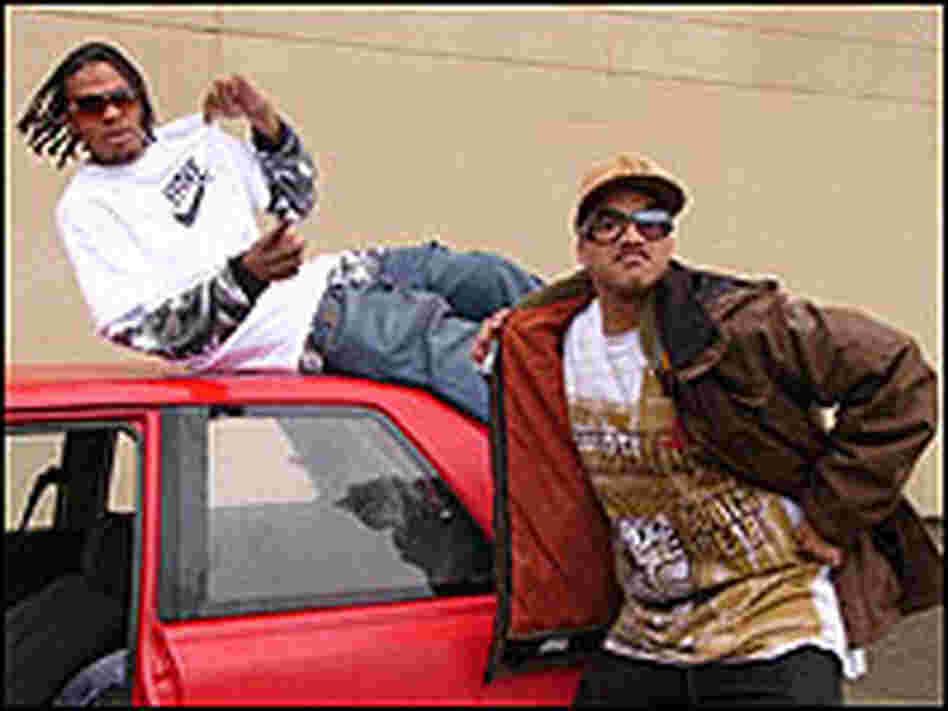 Two young men goof around on a battered red car, one wearing a Hyphy T-shirt.