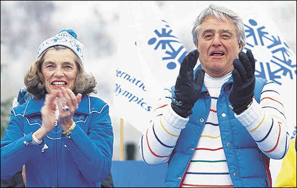 Shriver and her husband, wearing matching blue coats, applaud athletes at the Special Olympics.