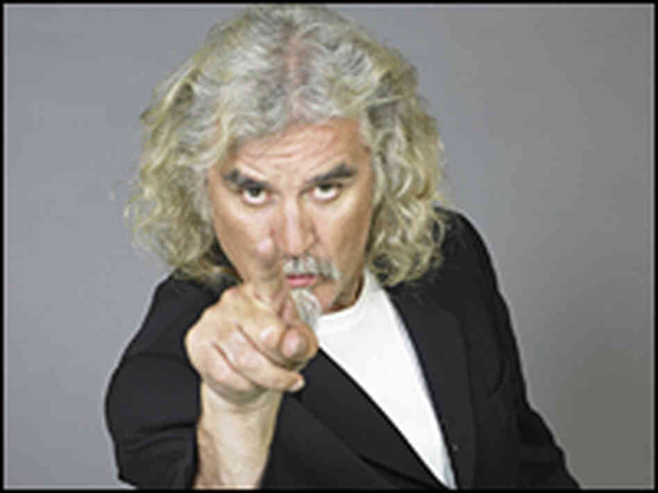 Billy Connolly, with shoulder-length white-gray hair and a goatee, points directly at the camera.