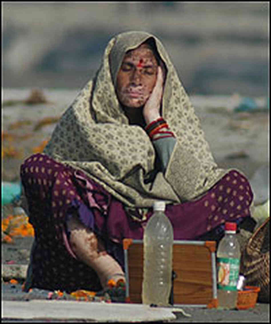 A woman with a skin disease rests near offerings and trash on the banks of the Ganges.