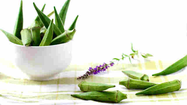 Fresh okra is piled into a white bowl and scattered on a checkered tablecloth