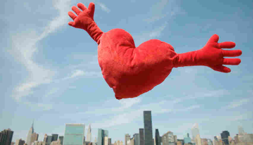 Red heart cushion with arms flies over city skyline with arms wide open for a big hug.