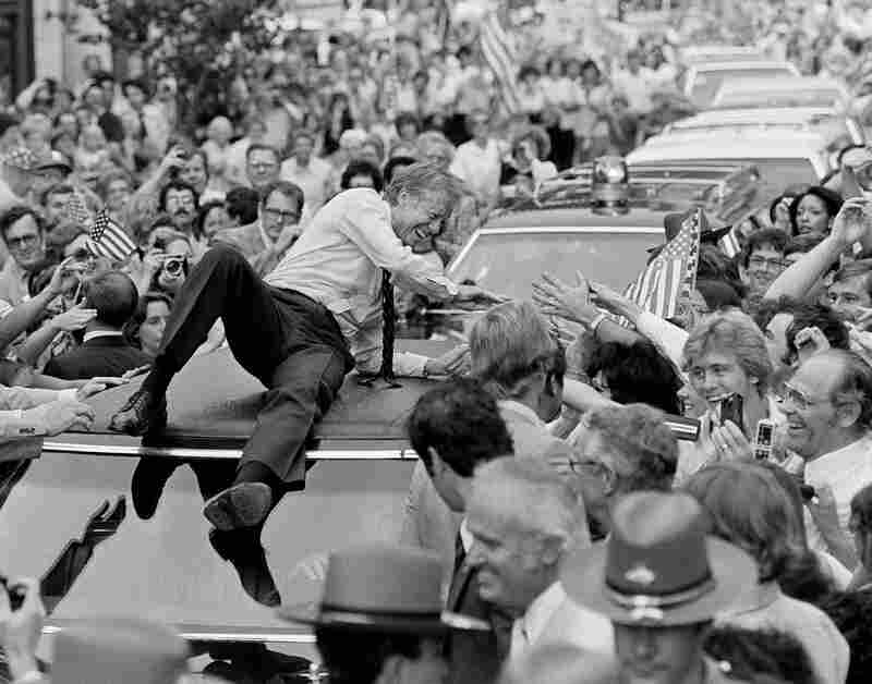President Carter leans across the roof of his car to shake hands along the parade route on his way to a town meeting in Bardstown, Ky., 1979.