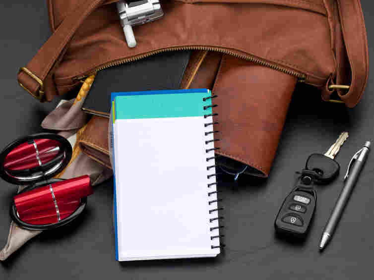 Personal organizer with blank lined page propped against various items spilling out of a brown leather purse. Studio isolated on a black textured background.