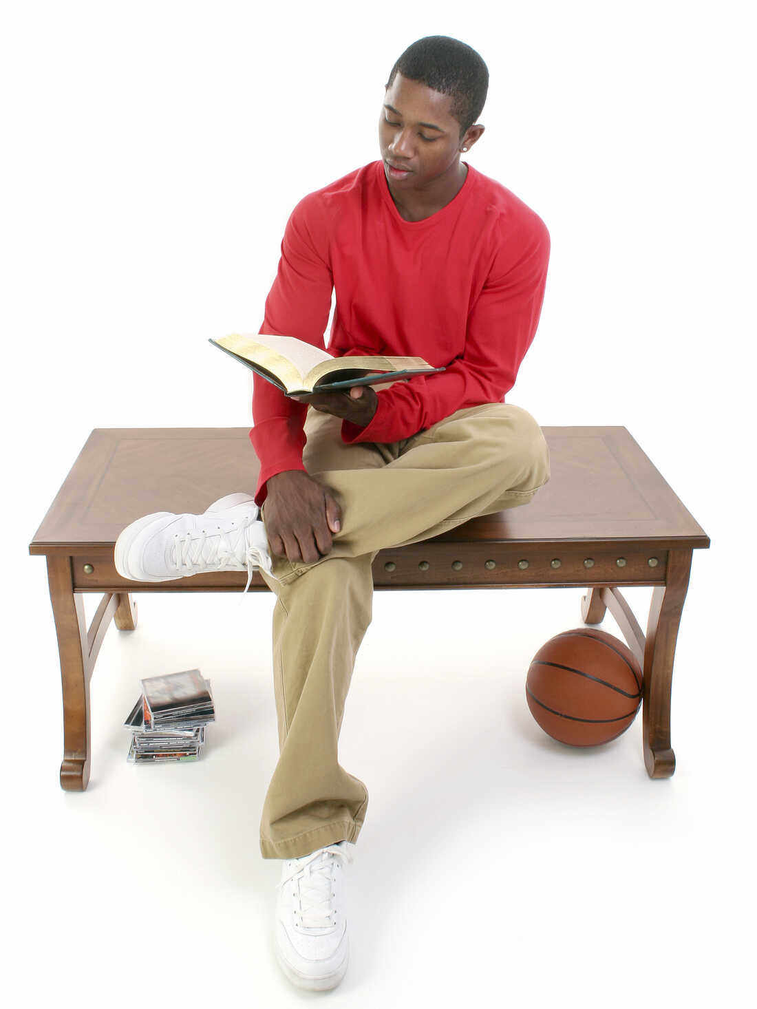 Casual man sitting on a table reading a book, with a stack of video games and a basketball nearby.