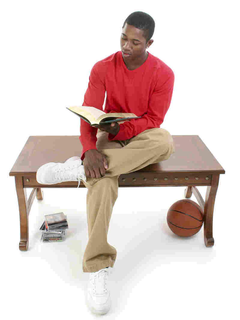 Casual man sitting on a table reading a book, with a stack of video games and a basketball nearby.