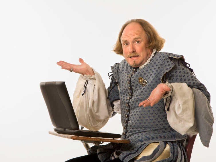 William Shakespeare in period clothing sitting in school desk with laptop computer shrugging at viewer.