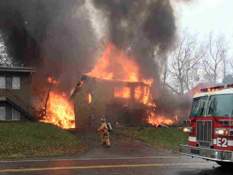 A firefighter walks up a driveway as an apartment building burns after being struck by a small business jet Tuesday. The crash killed nine people.