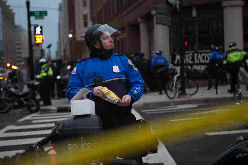 A police officer takes a break to eat a snack while protestors remain peacefully detained in the background.