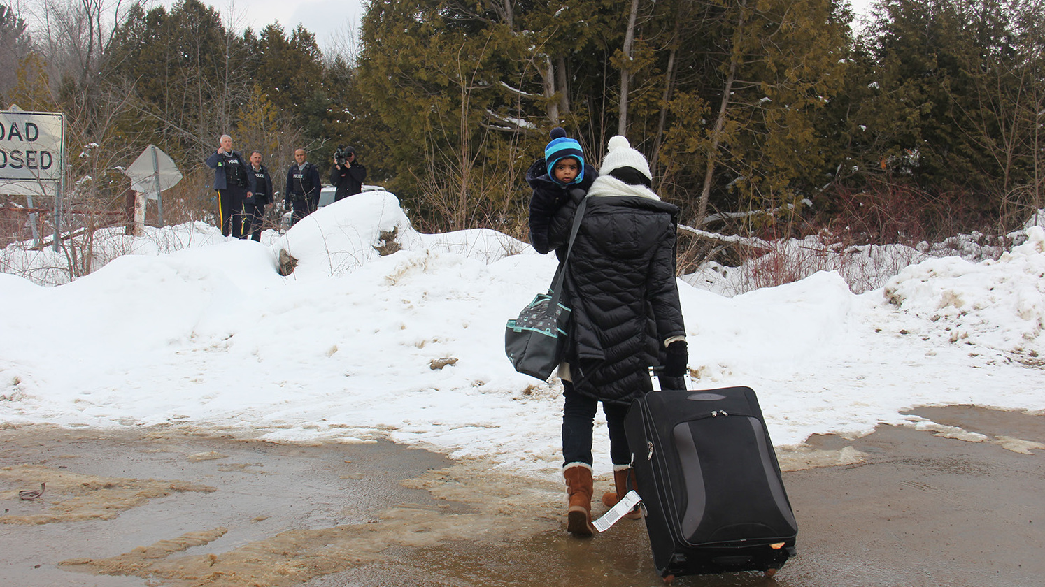 A woman traveling alone with her infant, seeming to understand that she will be arrested, walks toward Canadian police on the far side of the border. (VPR)