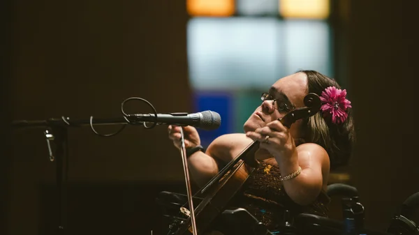 A woman with a flower in her hair plays the violin onstage in front of a microphone.
