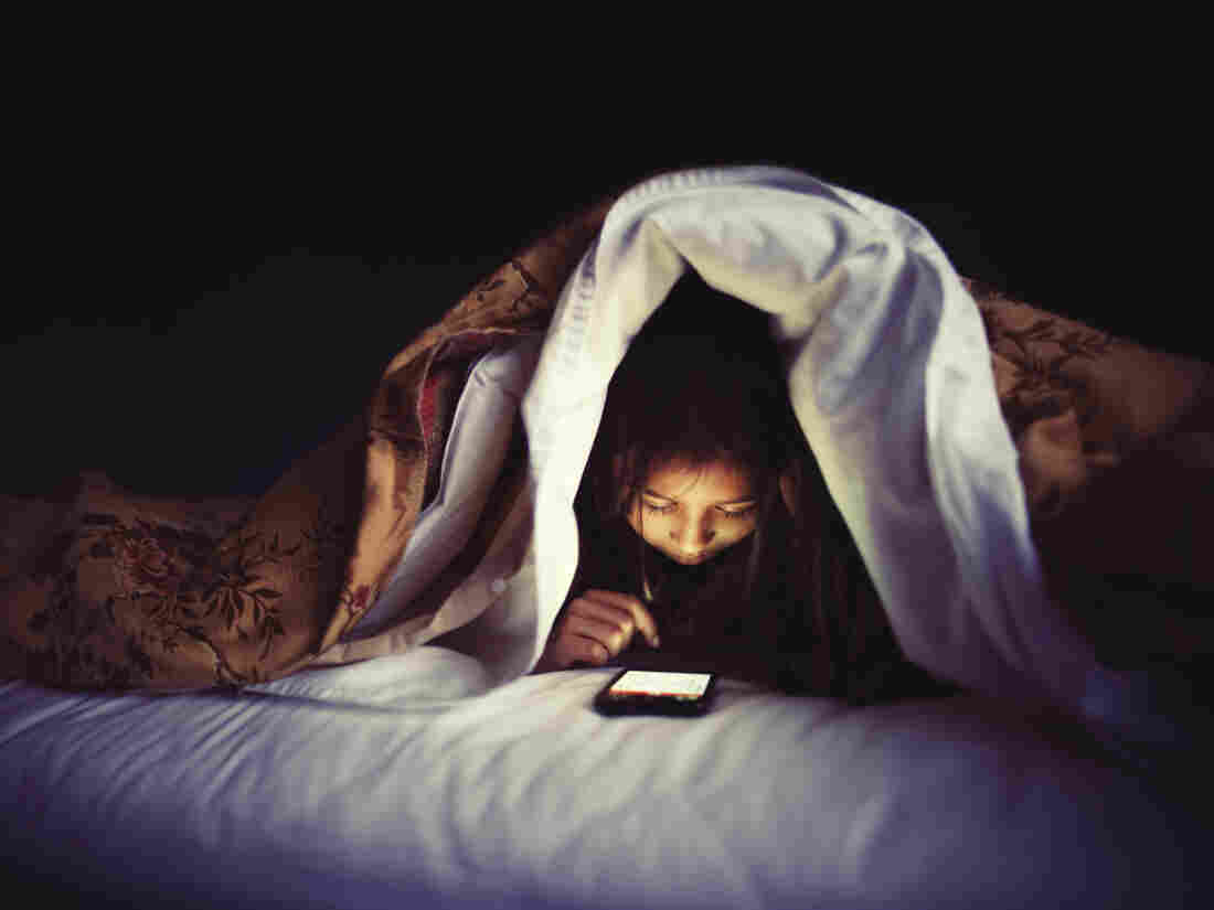 Girl using smartphone under bed covers.