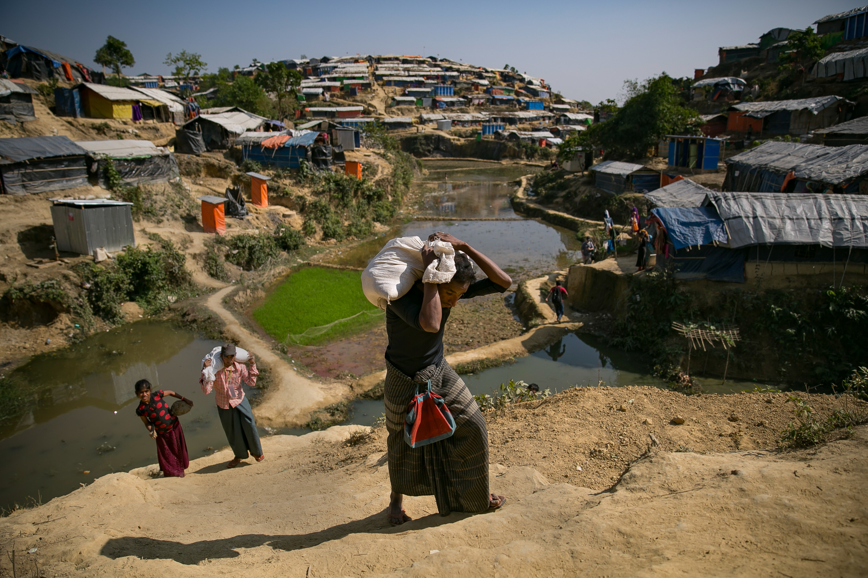 Mohamed Yonus (dark shirt, green skirt) carries his distribution bag to his home in the Hakimpara refugee camp. (Allison Joyce for NPR)