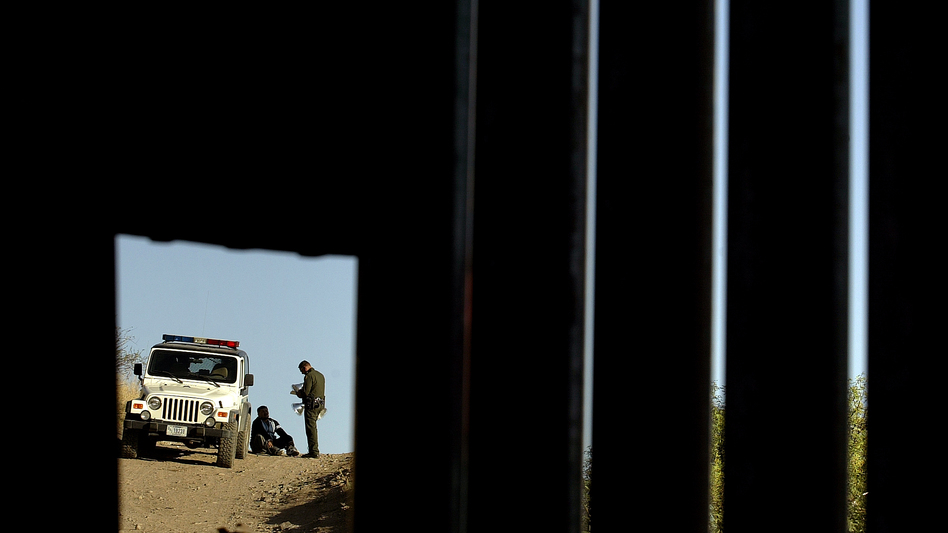 A U.S. Border Patrol agent questions a man in Nogales, Ariz., seen through a hole in a metal fence marking the border between the U.S. and Mexico, in 2007. (Guillermo Arias/Associated Press)
