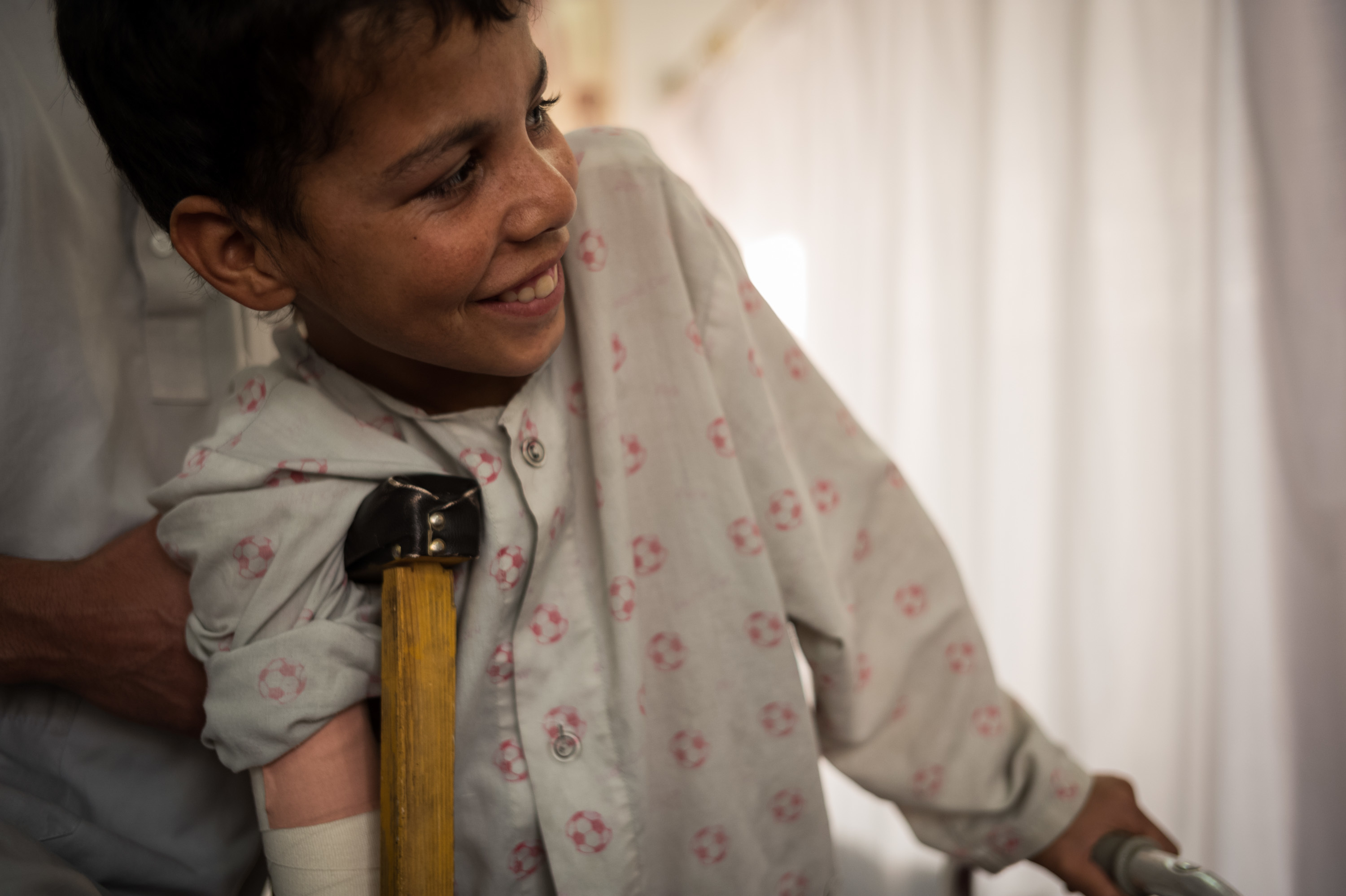 Barkatullah smiles and rests on a crutch and grips his walker during a physical therapy session at Emergency War and Trauma Hospital in Kabul. The 13-year-old lost his right arm and leg in an explosion. He practices standing on the walker 30 seconds at a time. (Ivan Armando Flores for NPR)