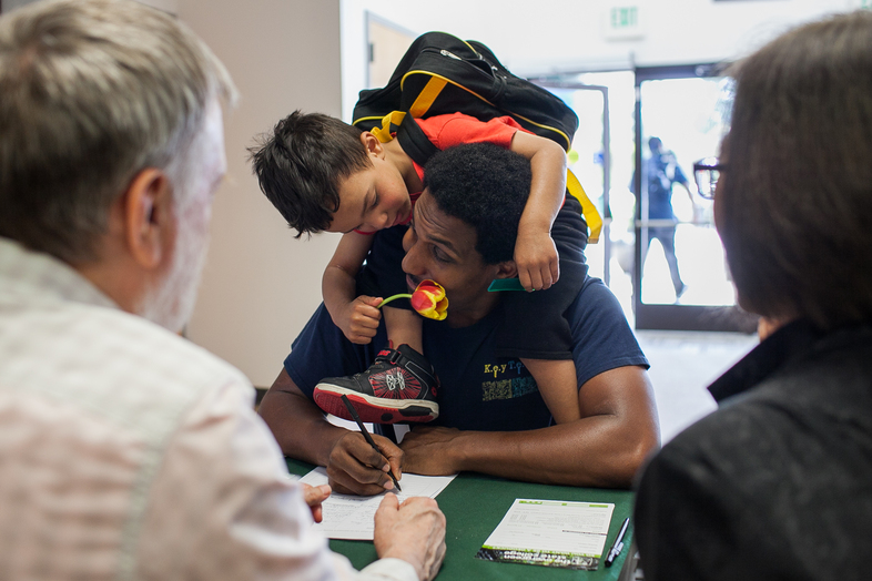 Nathan Powers, 3, offers his father, Adam Powers, a flower while he talks with campus representatives at the Spring Fair at Evergreen State College's Tacoma, Wash. campus on May 19. Powers, of Des Moines, Wash., works at a nonprofit and is considering going back to school. (Jovelle Tamayo for NPR)