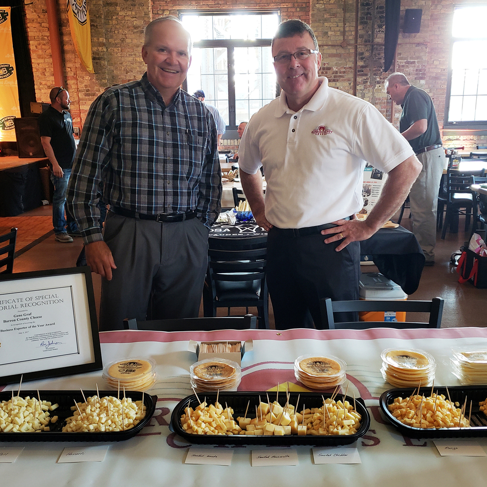 Gene Graf (right) and David Kochendorfer (left) of Barron County Cheese behind their cheese table at a Wisconsin event promoting cheese for export. (Maayan Silver/Milwaukee Public Radio)