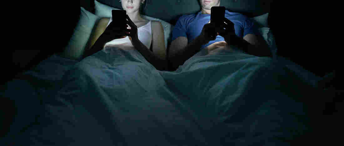A couple side by side in bed, each looking at their cell phones.