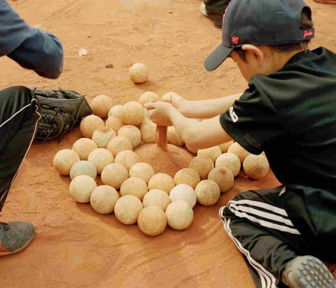 Children play in the sand of a baseball field next to a pile of baseballs.