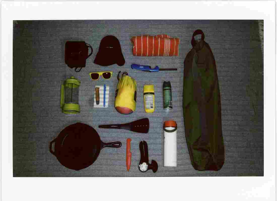 A polaroid photo of camping gear including a camping chair, sun screen, bug spray, sunglasses, a cast iron skillet, etc., arranged in a neat grid on a blue blanket.
