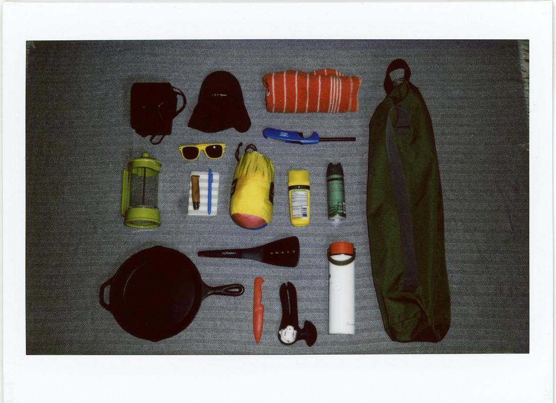 A polaroid photo of camping gear including a camping chair, sun screen, bug spray, sunglasses, a cast iron skillet, etc., arranged in a neat grid on a blue blanket.