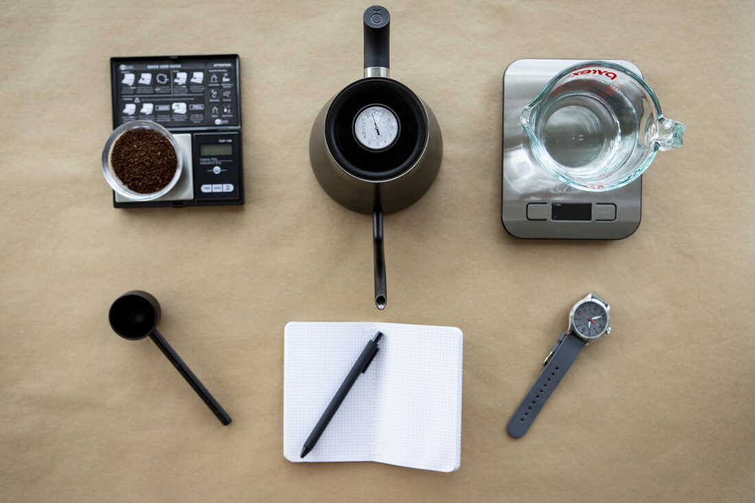 Coffee grounds on a scale, a black gooseneck kettle, water on a scale, a coffee scoop, a notebook and pen and a watch are photographed from above on a tan background.