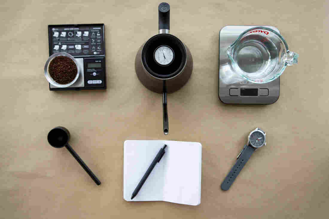 Coffee grounds on a scale, a black gooseneck kettle, water on a scale, a coffee scoop, a notebook and pen and a watch are photographed from above on a tan background.