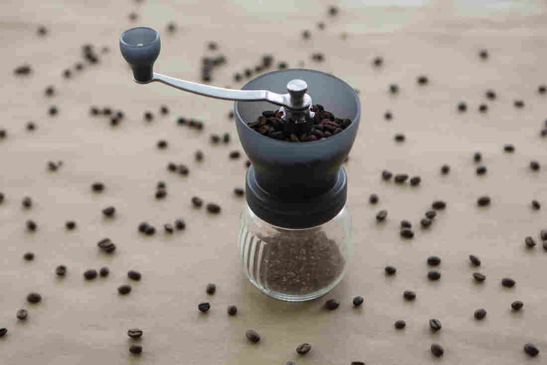 A manual burr grinder filled with beans is photographed on top of tan paper scattered with coffee beans.