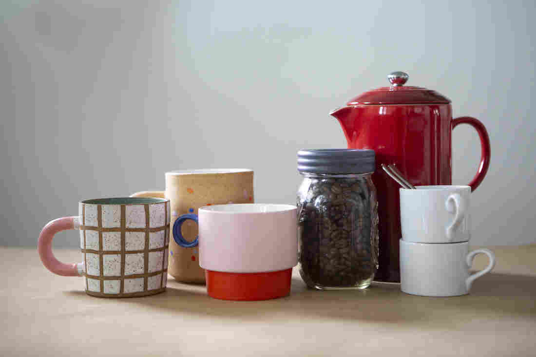 Three coffee mugs, a red ceramic french press, a glass jar of beans and two white espresso cups are grouped on a tan table with a white background.