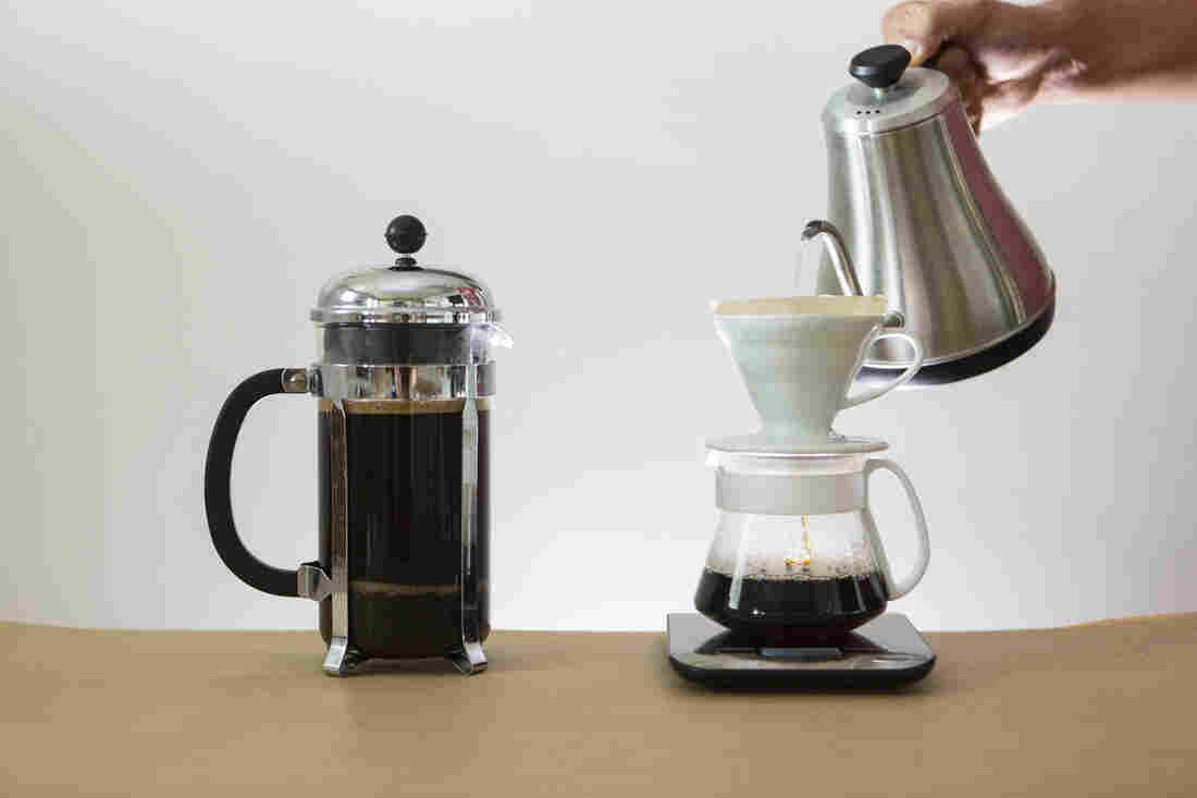 A french press full of coffee on the left and a pour over device with a hand pouring water over coffee sit on a tan table top against a white backdrop.
