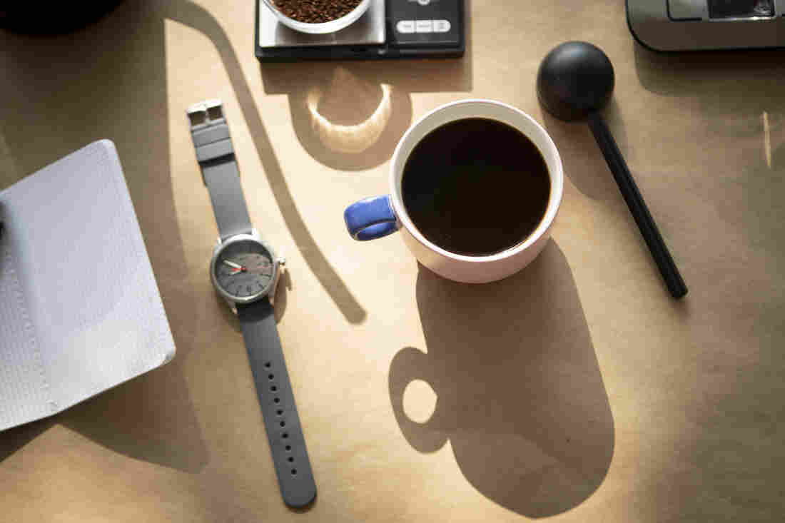 A mug full of coffee, a watch, a coffee scoop, a notebook, a scale and the shadow of a goose neck kettle are photographed from above against a tan backdrop.