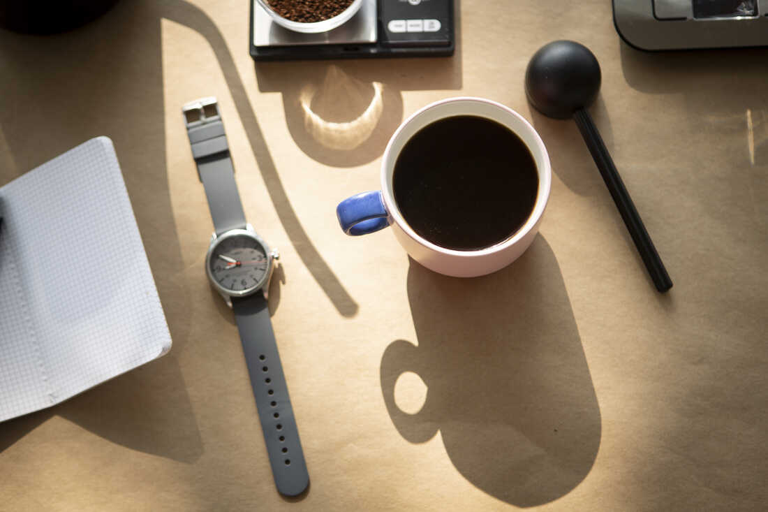 A mug full of coffee, a watch, a coffee scoop, a notebook, a scale and the shadow of a goose neck kettle are photographed from above against a tan backdrop.